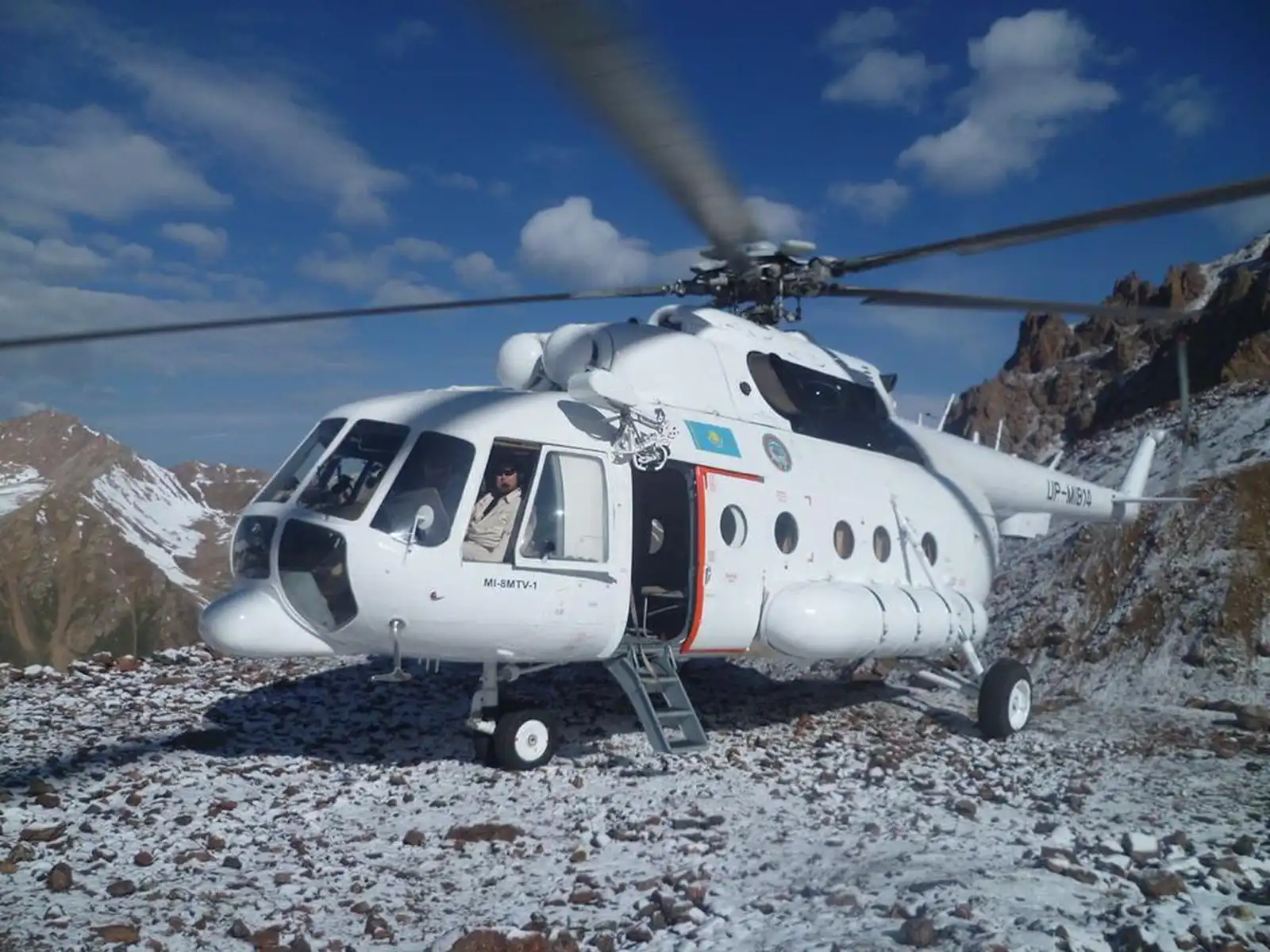 Mi-8 helicopter landed on a rocky snow-covered mountain slope with pilot visible in the cabin